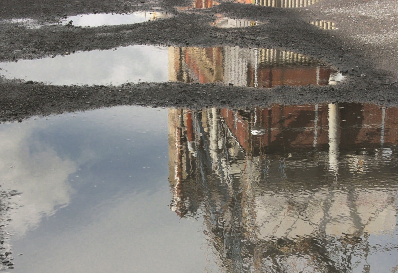 The picture shows two puddles on a gravel surface in which an industrial building is reflected.