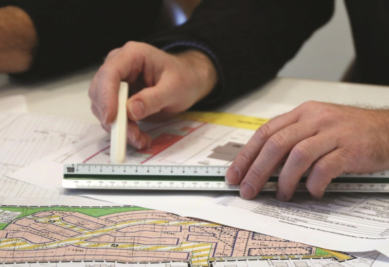 The picture shows two hands with a ruler and a pencil on a development plan spread out on the table.