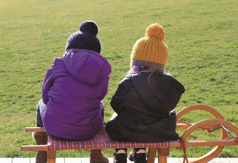 The picture shows two children from behind, wearing bobble hats and winter jackets, sitting next to each other on a sledge that is standing on a wooden terrace in front of a green meadow.