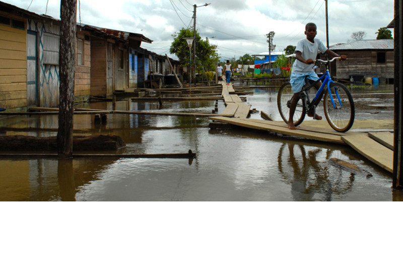 The picture shows a young dark-skinned woman from behind, riding a bicycle over a footbridge flooded with water. The bicycle is heavily loaded with a large sack and firewood.