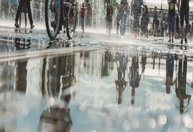 Das Bild zeigt eine Wasserfläche in eine Stadt, offensichtlich ein Brunnen. Es laufen mehrere Menschen über die Wasserfläche und spiegeln sich darin.