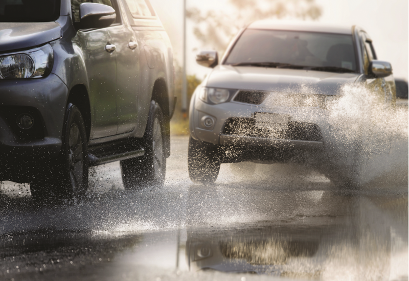 The picture shows a close-up of a roadway covered with water. A car is almost hitting another car.