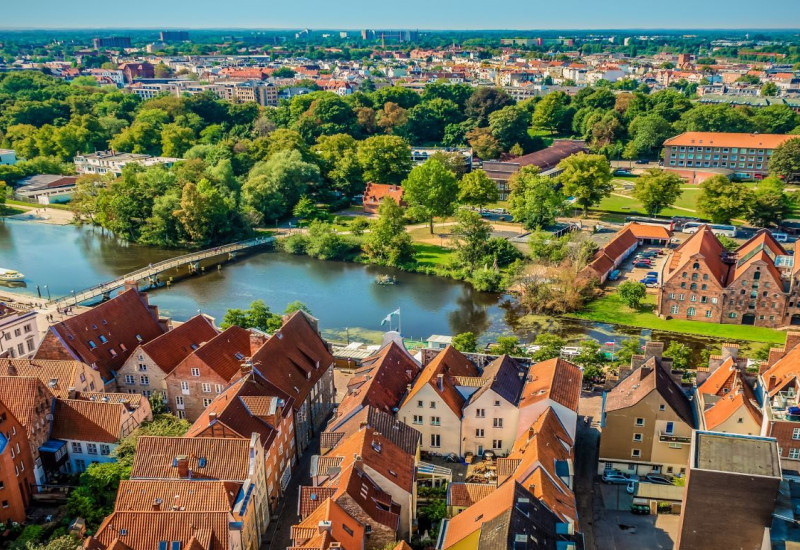 Der Blick geht von oben in die Ferne über eine Stadt mit roten Ziegelsteindächern. Die Stadt ist umgeben von Grün und liegt an einem Fluss. Der Himmel ist blau.