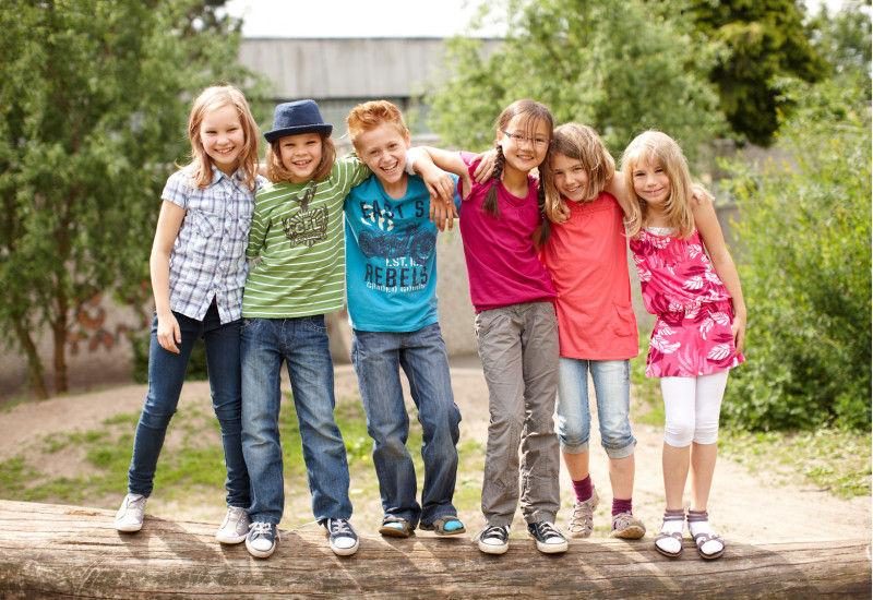 6 Kinder stehen lachend auf einem liegenden Baumstamm für ein Gruppenfoto, die Arme jeweils auf den Schultern des Nachbarn