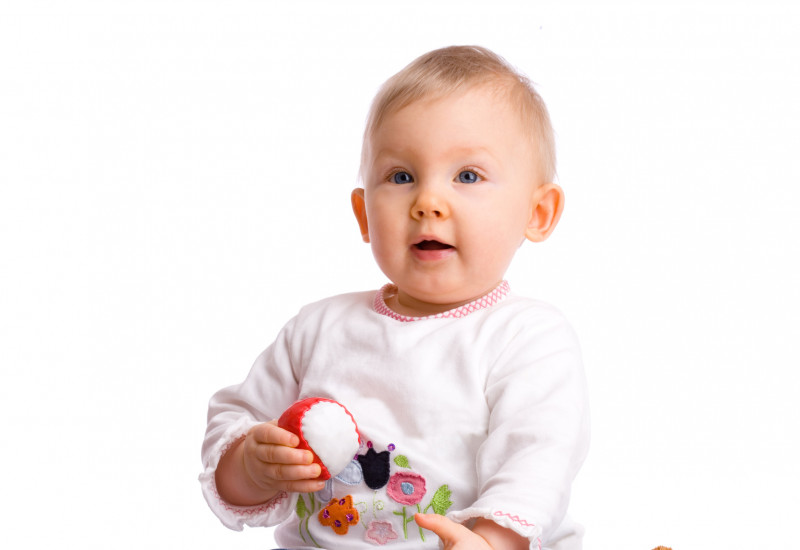 A baby sits among cuddly toys with a ball in its hand.