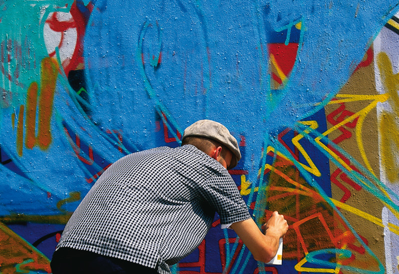 Mann der gebeugt vor einer Wand steht und ein bereits existierendes Graffitti übersprüht