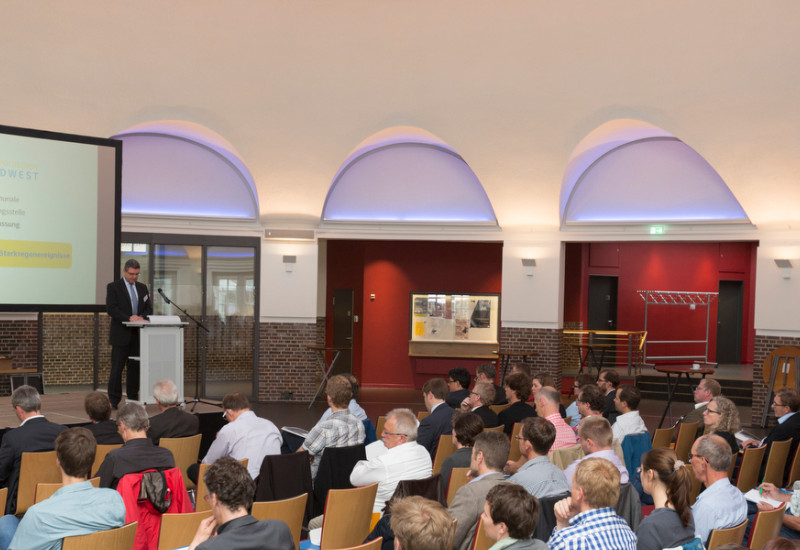 Viele Personen sitzen vor einer Bühne und hören einem Redner zu