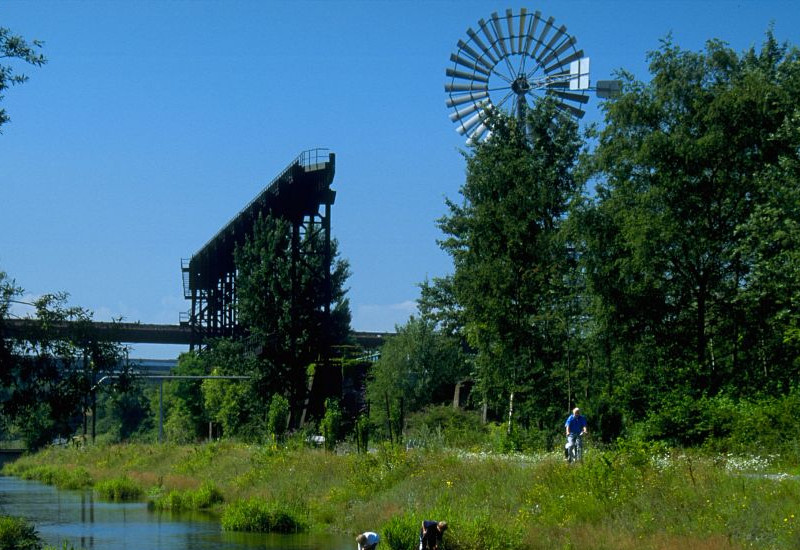 Ein Gewässer in einem Landschaftspark. Im Hintergrund steht ein Windrad. Im Vordergrund spielen Kinder in den Uferbereichen und fangen Fische.