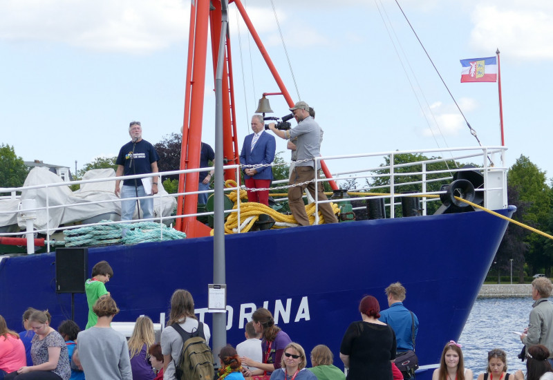 Menschenmenge vor einem Schiff, auf dem Redner und Kameramann stehen.