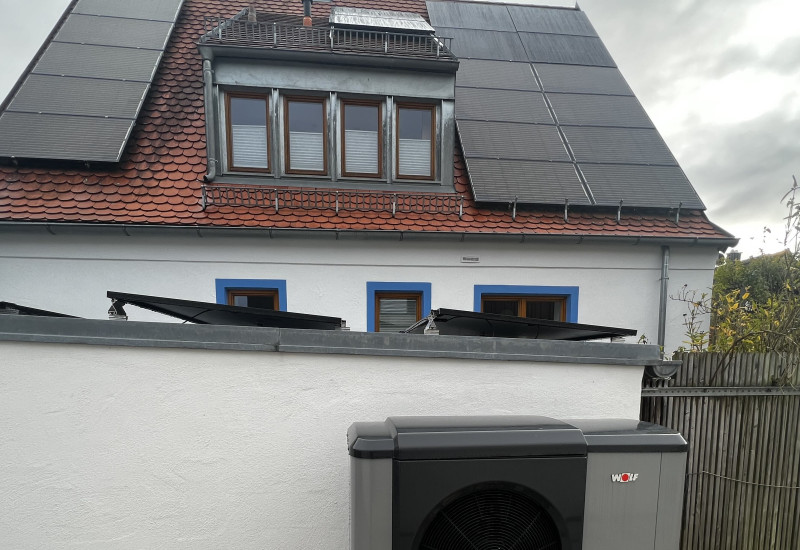 Eine Wärmepumpe an einer Grundstücksmauer, im Hintergrund das Einfamilienhaus.