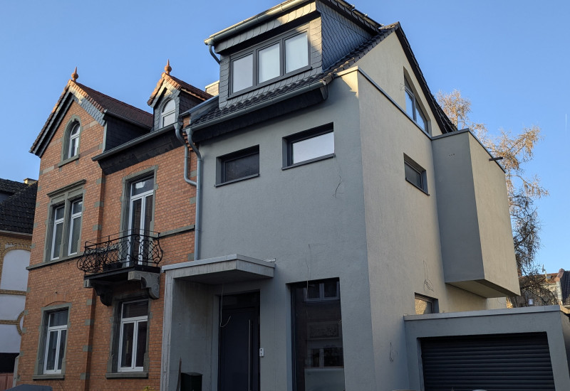 Die Fassade eines zweigeschossigen, rot geklinkerten Altbaus mit einem modernen, grau verputzten Anbau.