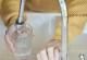Nahaufnahme einer Person in einem gelben Pullover, die ein Glas unter einem Wasserhahn füllt. Das Wasser läuft in das geriffelte Glas. Im Vordergrund ist der Chrom-Wasserhahn zu sehen.