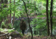 Fotografie eines Menschen, der mit Naturmaterialien verkleidet ist und im Wald vor einem Teich steht