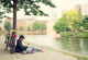 Ein junger Mann sitzt in der Stadt an einem Fluss unter einem Baum und schaut auf sein Tablet mit Kopfhörern im Ohr. Am Baum lehnt sein Fahrrad.