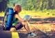 eine junge, sportlich gekleidete Frau mit Rucksack, Strohhut und Isomatte wartet auf der Bahnsteigkante in der Sonne sitzend auf einen Zug und schaut in eine Landkarte