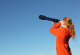 eine junge Frau schaut mit einem Fernrohr in die Ferne, im Hintergrund klarer, blauer Himmel