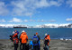 Reisende stehen im Vordergrund auf Land. Im Hintergrund sieht man antarktisches Meer und ein großes Schiff vor beschneiten Hügeln. 