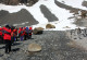 Auf der linken Seite des Bildes steht eine Gruppe von Menschen in roten Kälteschutzanzügen. Sie halten ausreichend Abstand zu einer Gruppe Pinguine auf der rechten Seite. 