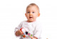 Ein Baby sitzt neben Kuscheltieren, es hält einen Ball in der Hand und guckt erwartungsvoll nach oben.