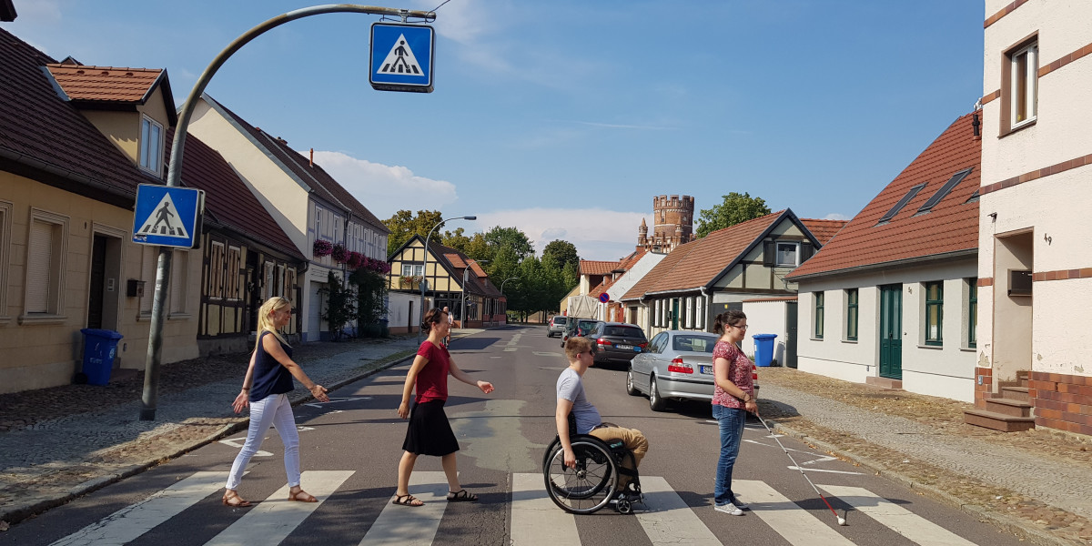 Straße mit niedrigen Fachwerkhäusern und einem Zebrastreifen, der von einer Frau mit Blindenstock, einem Rollstuhlfahrer und zwei weiteren Fußgängerinnen überquert wird.