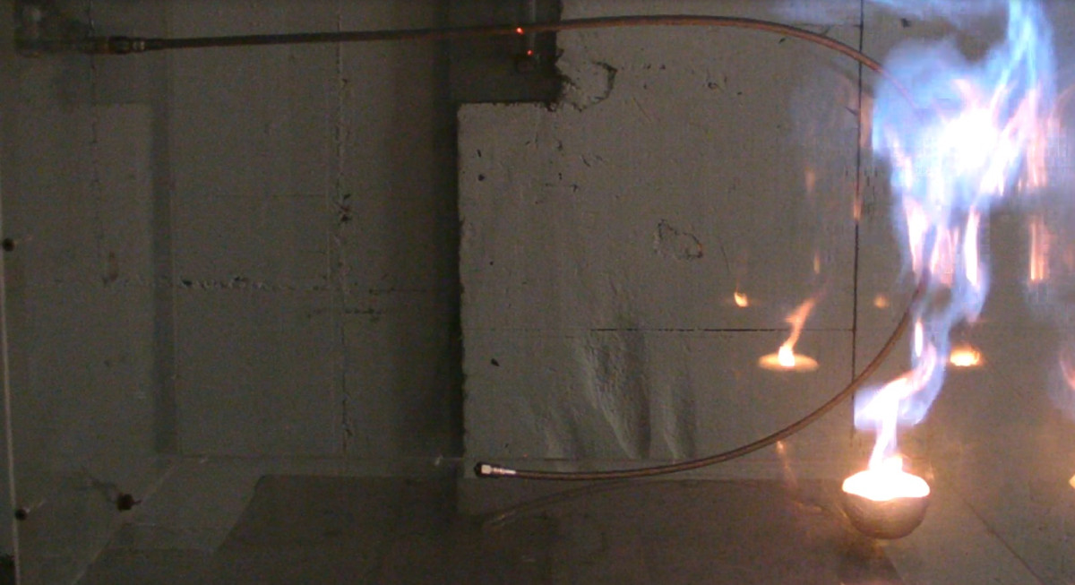 Ein Metallrohr ist U-förmig vor einer Wand gebogen. Auf der rechten Seite lodert eine große Flamme das ganze Rohr entlang.