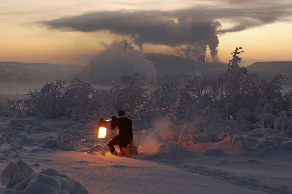 Mann mit leuchtender Laterne in einer eisigen Schneelandschaft. Im Hintergrund rauchende Industrieschornsteine.