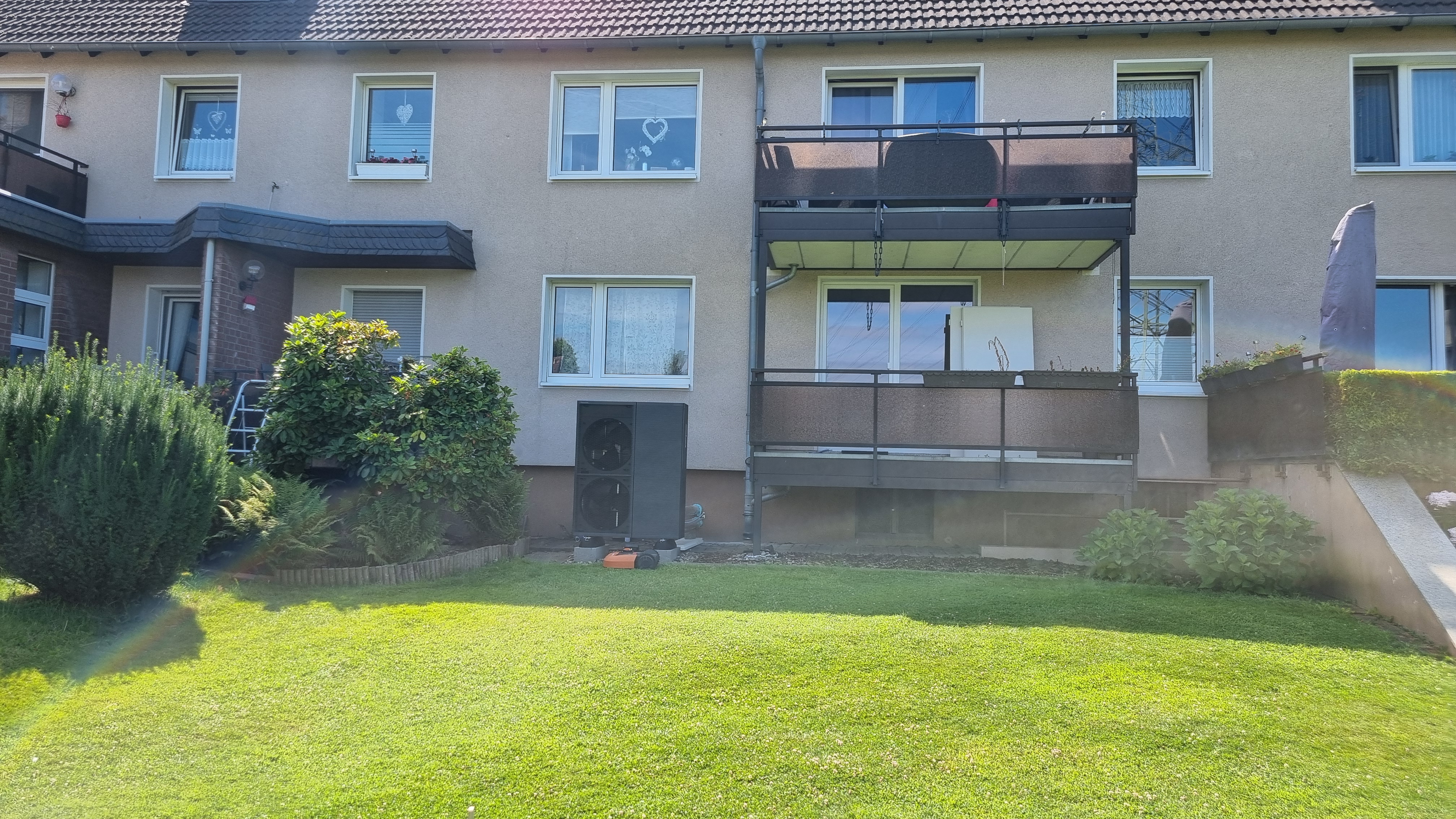 Ansicht eines zweistöckigen Mehrfamilienhauses mit Rasenfläche und einzelnen Büschen davor. Vor dem Haus steht das dunkelgraue Außengerät der Luft-Wasser-Wärmepumpe