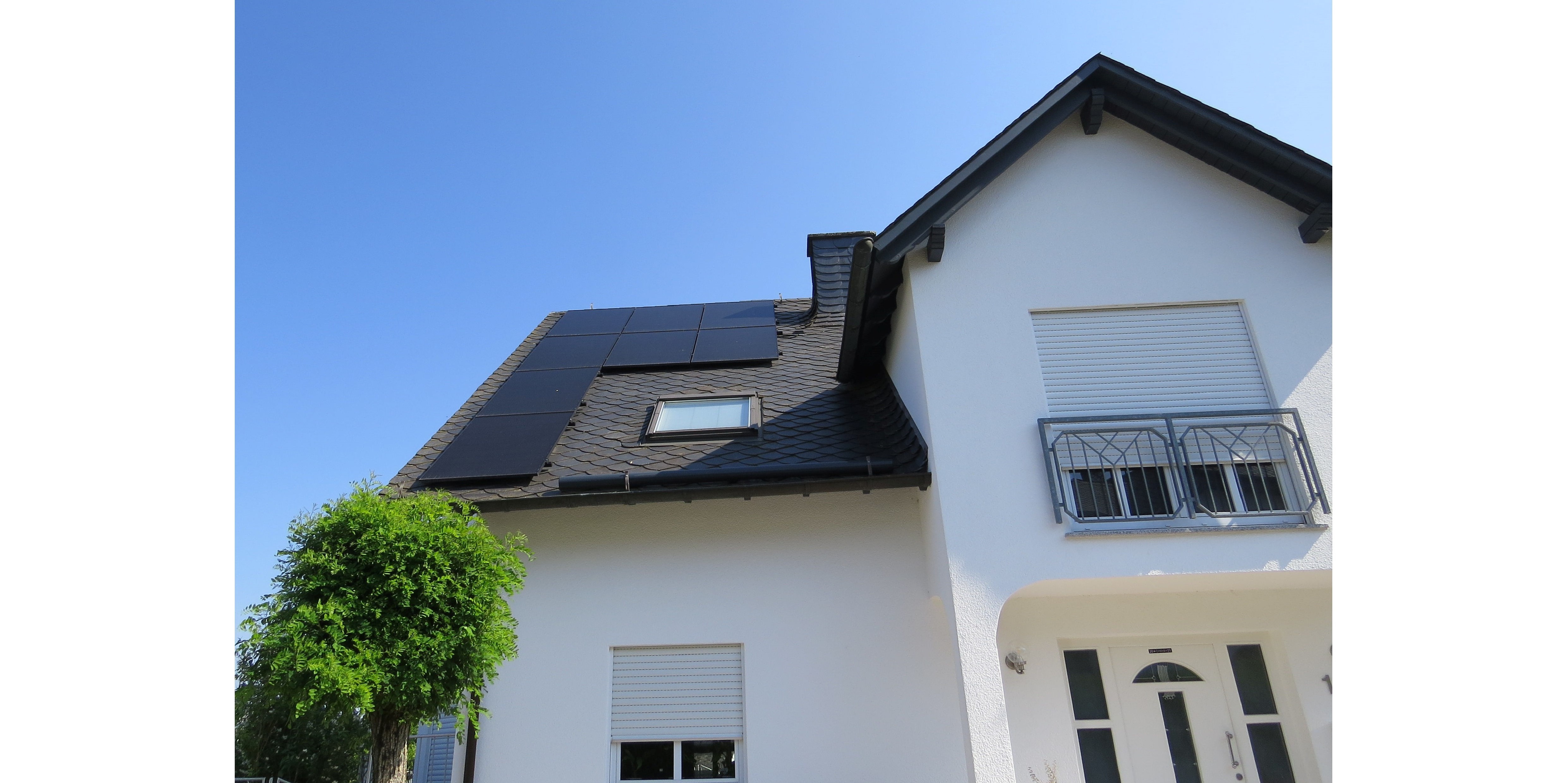 Ein weißes Einfamilienhaus mit großem Erker und PV auf dem schwarzen Dach vor blauem Himmel..