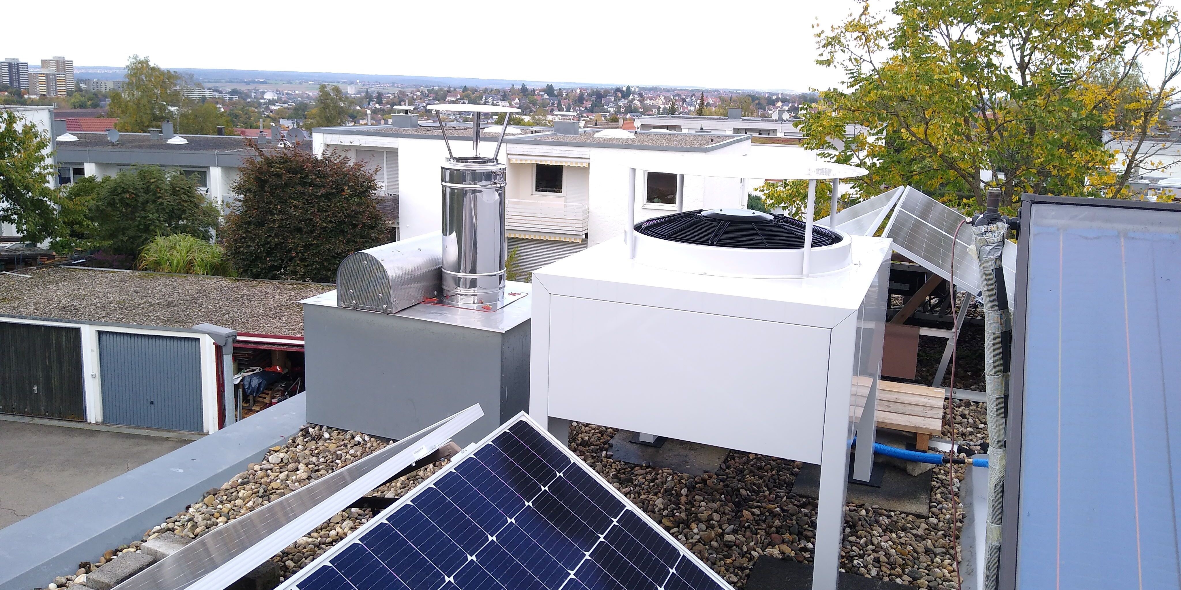 Die weiße Außeneinheit der Wärmepumpe steht am Rand des geschotterten Dachs; im Vorgergrund sind Photovoltaikmodule und am Bildrand ein Solarkollektor zu sehen.