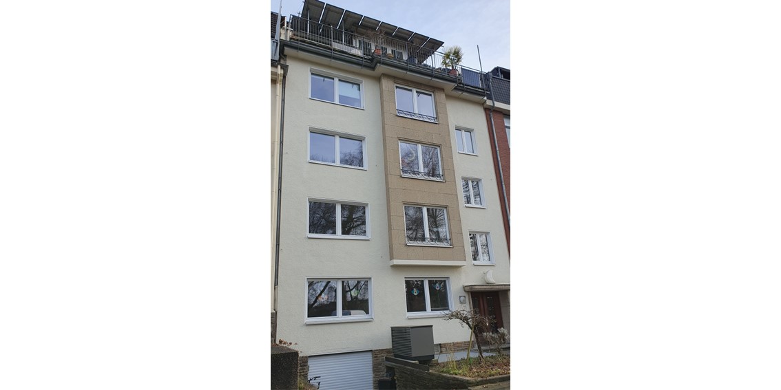 Die Gesamtansicht eines fünfstöckigen hell-beigen Mehrfamilienhauses; über drei Stockwerke ist ein flacher Erker etwas dunkler abgehoben. An der Hauswand steht die Wärmepumpe, die nicht bis zur untersten Fensterkante reicht.