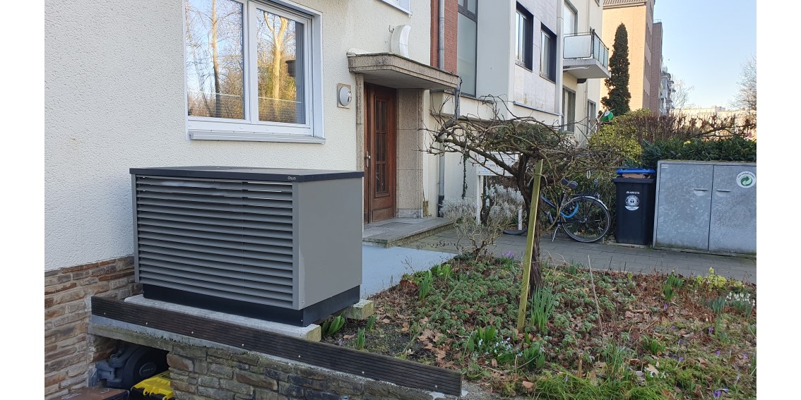Die Wärmepumpe im Vorgarten eines Mehrfamilienhauses. Über der Wärmepumpe ist ein Doppelfenster zu sehen sowie die Eingangstür.