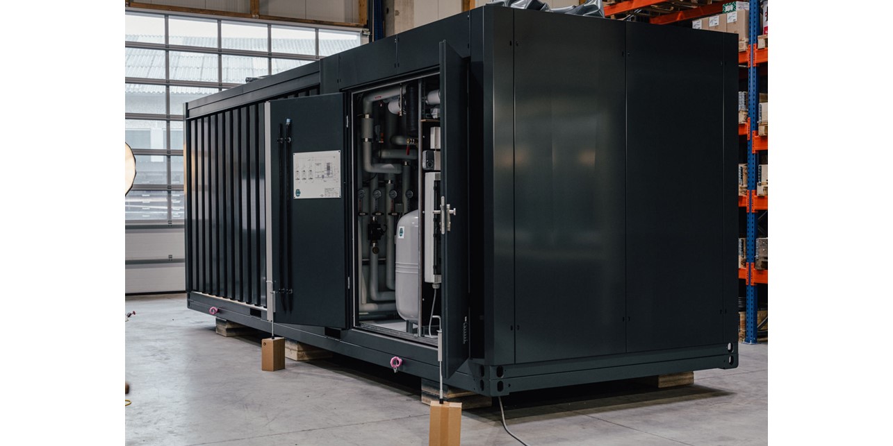 Die containerartige Wärmepumpenzentrale in einer Lagerhalle in Nahansicht; eine Tür ist offen, die einen Blick auf isolierte Rohre bietet.