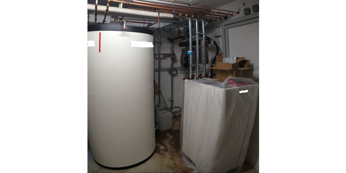 Die weiße Wärmepumpe im Heizraum am rechten Bildrand, in Folie eingepackt. Die Rohrleitungen sind noch nicht isoliert. Daneben steht der fast raumhohe Pufferspeicher