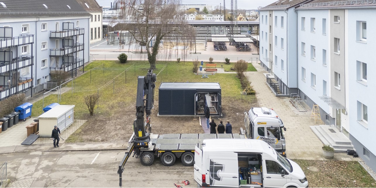 Aufstellung der containterartigen Wärmepumpenzentrale im Innenhof mit Autokran; links und rechts stehen zwei dreistöckige, hellgraue Mehrfamilienhäuser.