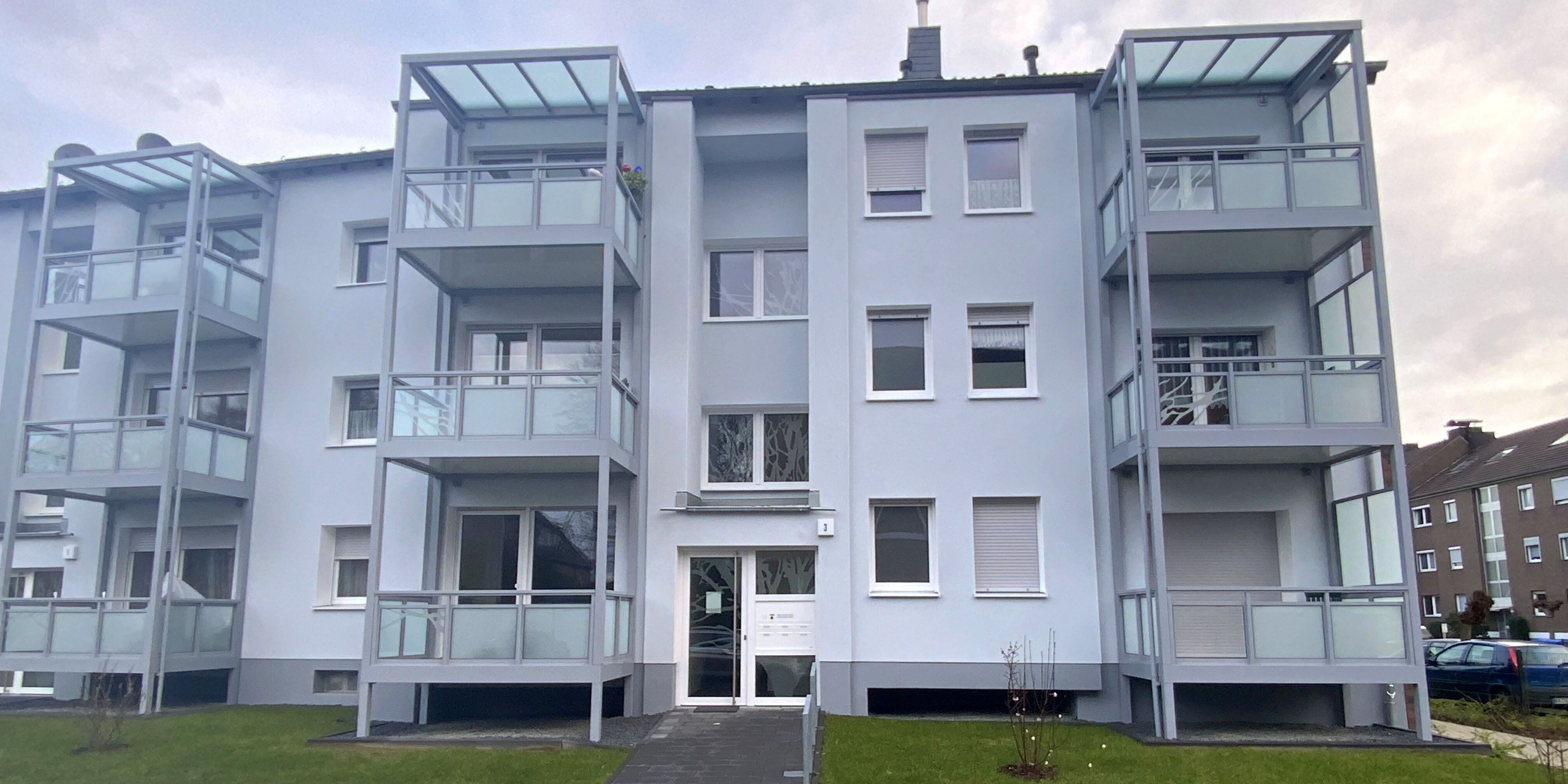 Ein dreistöckges, hellgraues Mehrfamilienhaus mit links und rechts vom Eingang vorangestellten Balkonen, frontal von vorne gesehen