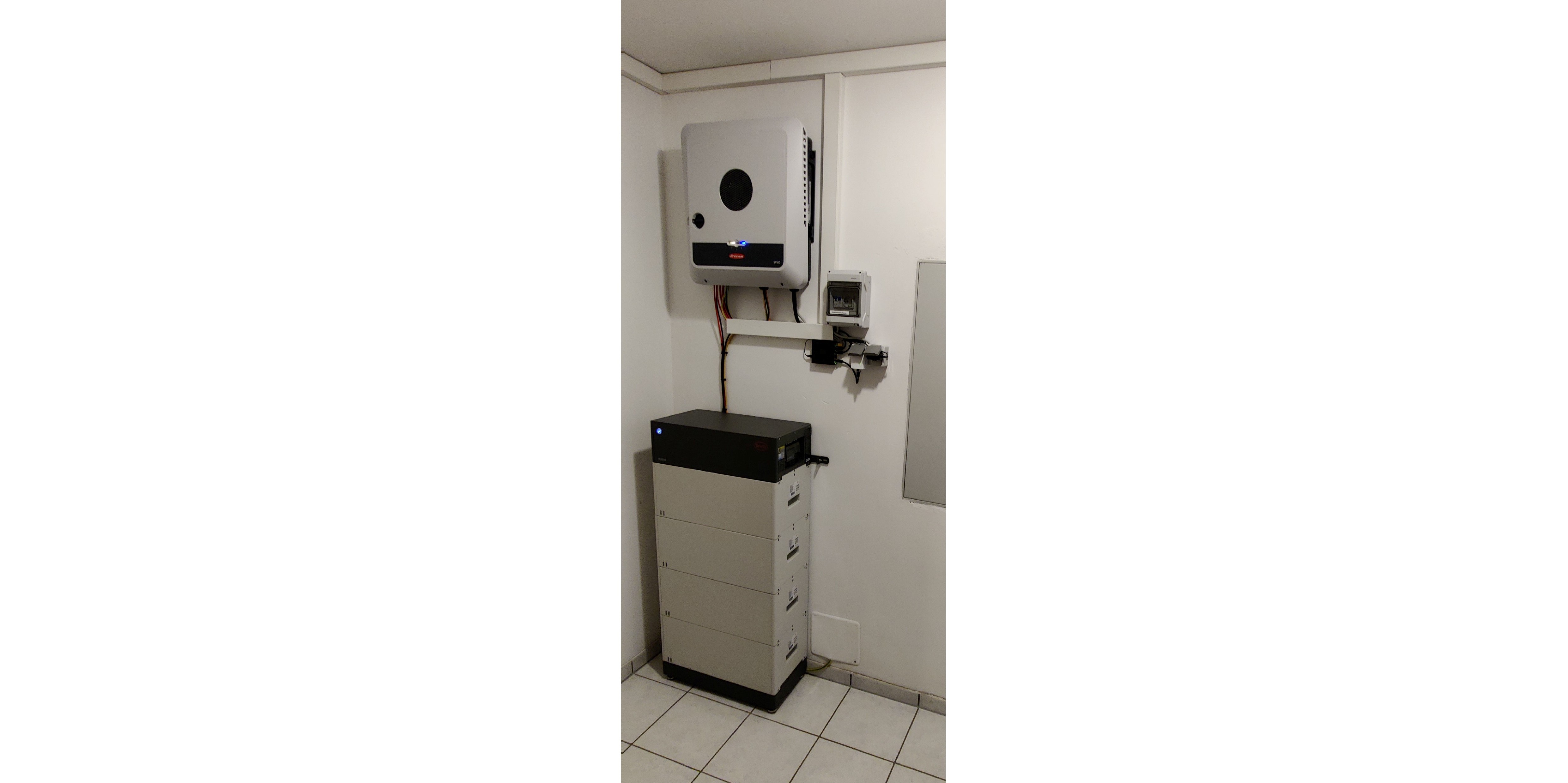An der weißen Kellerwand hängender Wechselrichter. Auf dem Boden davor steht in der Ecke der Batteriestapel.