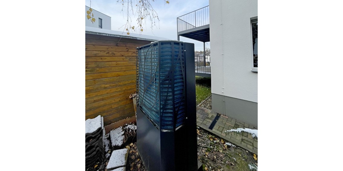 Das dunkelgraue Außengerät der Wärmepumpe im Garten zwischen Haus und Holzschuppen.