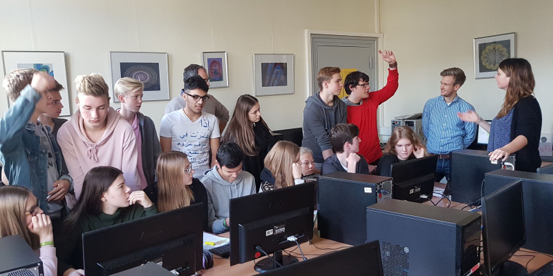 Lernende sitzen oder stehen im Klassenraum vor den Tischen mit Computern. Ein Schüler in einem roten Pullover meldet sich zu den gestikulierenden Ausführungen der Lehrenden.