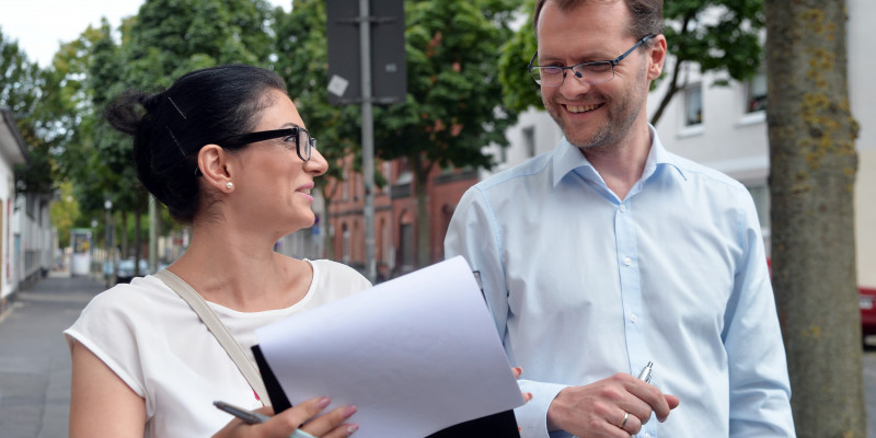 Ein Mann und eine Frau stehen sich unterhaltend und lachend nebeneinander. Die Frau hält ein Papier auf Brusthöhe.