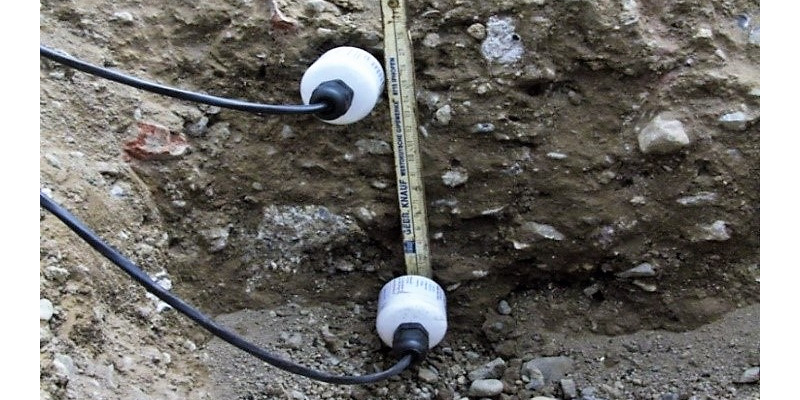 Ein Temperaturmessgerät in Röhrchenform liegt auf einen Bodenquerschnitt. Die weißrunden Tempertaturfühler liegen ebenfalls auf dem Bodenquerschnitt