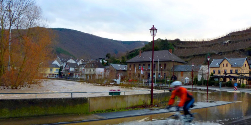 Person fährt mit dem Fahrrad durch Hochwasser in einer Ortschaft. Im Hintergrund sind Häuser und Berge zu sehen. Himmel ist bewölkt.