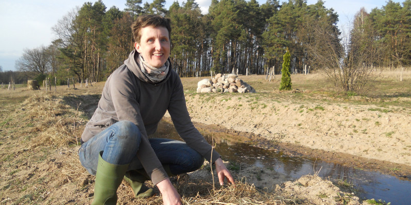 Eine Person in Gummistiefeln hockt neben dem Wassergraben.
