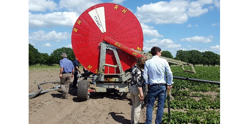 Exkursionsteilnehmende betrachten eine landwirtschaftlich genutzte Beregnungsfläche. Das Beregnungsgerät erscheint als große runde rotes Gebilde im Hintergrund vor wolkigem Himmel.