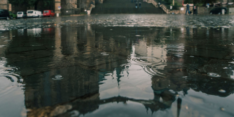Regentropfen fallen auf eine Wasserfläche. Im Hintergrund stehen parkende Autos im Wasser vor Gebäuden.