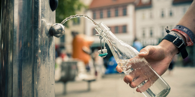 Eine Hand schöpft mithilfe einer wiederverwendbaren Glasflasche Wasser aus einem Trinkbrunnen in der Stadt.