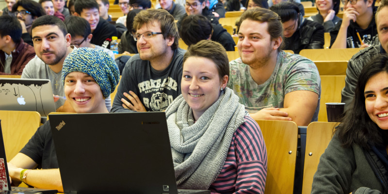 In einem Hörsaal sitzen Studierende, manche mit aufgeklapptem Laptop vor sich. Zwei Personen in der ersten Reihe schauen in die Kamera, andere schauen nach vorne und lachen.
