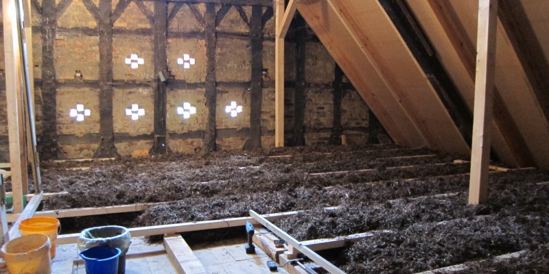 Seegras liegt zum Trocknen breit ausgelegt auf rechteckigen Holzlattengestellen auf dem Boden eines Dachbodens. Die Wände des Dachbodens bestehen aus Fachwerk und haben Öffnungen nach draussen.