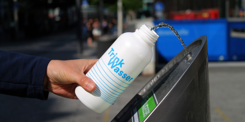 Eine Hand hält eine weisse Trinkflasche mit einem hellblauen Schriftzug: Trink Wasser.