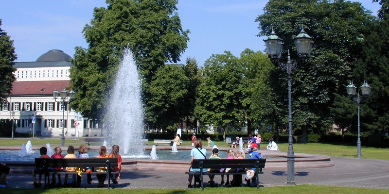 Eine große und einige kleinere Wasserfontänen entspringen einem Brunnen auf einem runden öffentlichen Platz. Drumherum sitzen Personen auf Bänken. Im  Hintergrund stehen hohe begrünte Bäume vor Gebäude und blauem Himmel.