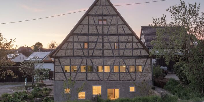 Foto der Giebelaußenwand einer Fachwerkscheune. Das Erdgeschoss ist aus Sichtbeton mit fünf versetzt angeordnet, quadratischen Fenstern unterschiedlicher Größen. Abenddämmerung. In den Fenstern warmes Licht.